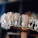 A sparkling silver tiara featuring intricate heart-shaped designs and dangling teardrop zirconia stones. The large zirconia stones are suspended to create a dynamic, shimmering effect.