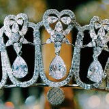 A sparkling silver tiara featuring intricate heart-shaped designs and dangling teardrop zirconia stones. The large zirconia stones are suspended to create a dynamic, shimmering effect.