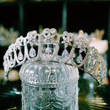  sparkling silver tiara featuring intricate heart-shaped designs and dangling teardrop zirconia stones. The large zirconia stones are suspended to create a dynamic, shimmering effect.